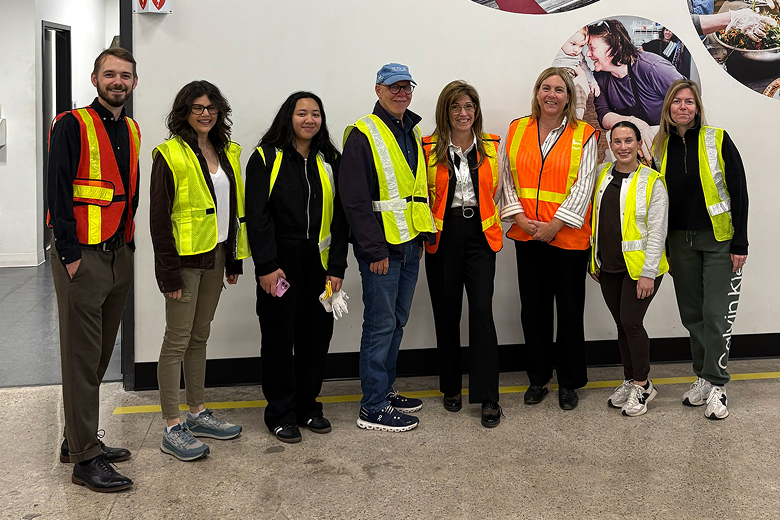 Group of eight people standing indoors against a white wall, wearing high-visibility safety vests in yellow and orange, some smiling at the camera.
