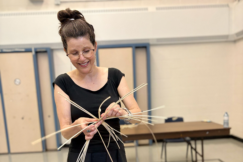 Woman with glasses and hair in a bun weaving thin wooden strips indoors.