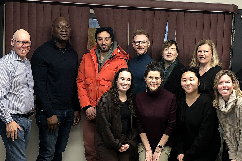 Group photo of ten adults smiling indoors in front of closed brown blinds.