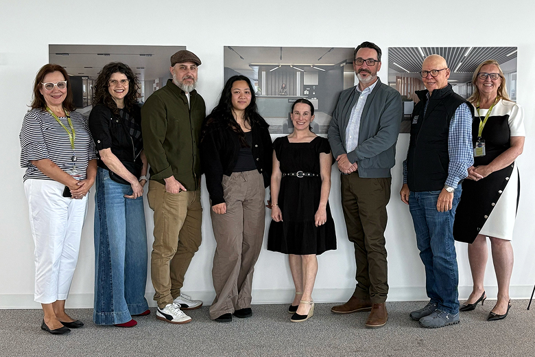 Eight people standing side by side indoors against a white wall with framed architectural photos behind them.