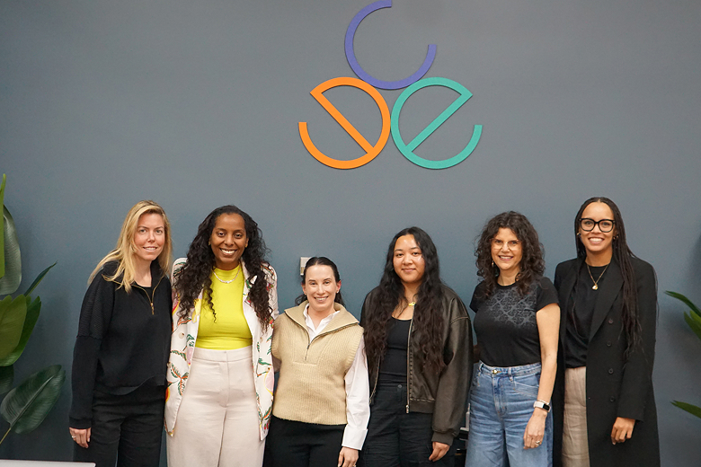 Six diverse women standing side by side in front of a gray wall with a colorful abstract logo above them.