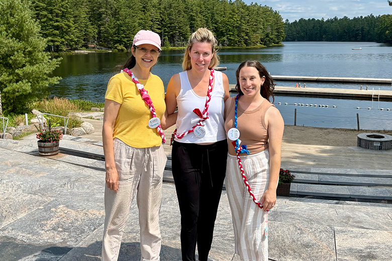 Three women standing on stone steps near a lake with docks and surrounded by trees on a sunny day.