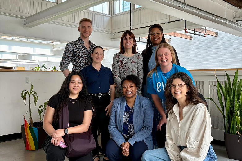 Group of eight diverse smiling people posing indoors in casual clothing with plants in the background.