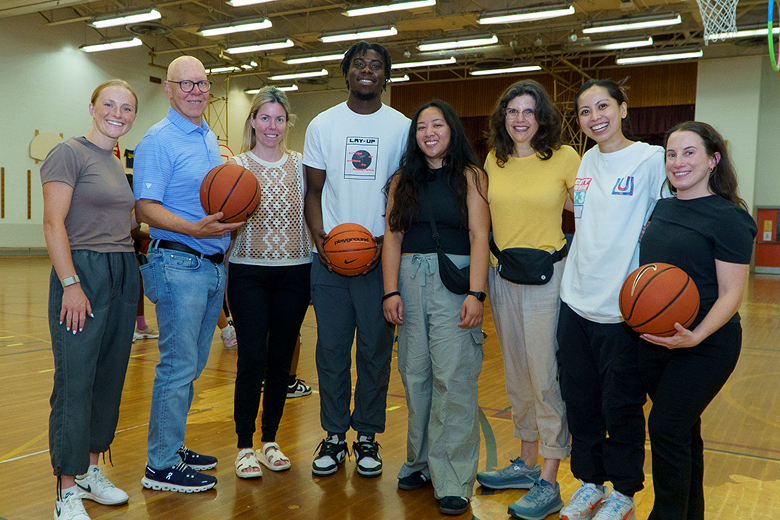 Group of eight diverse adults standing in a gymnasium, some holding basketballs, smiling at the camera.