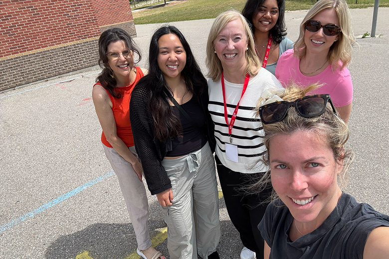 Group of six diverse women smiling outdoors on a sunny day, standing on pavement near a brick wall.