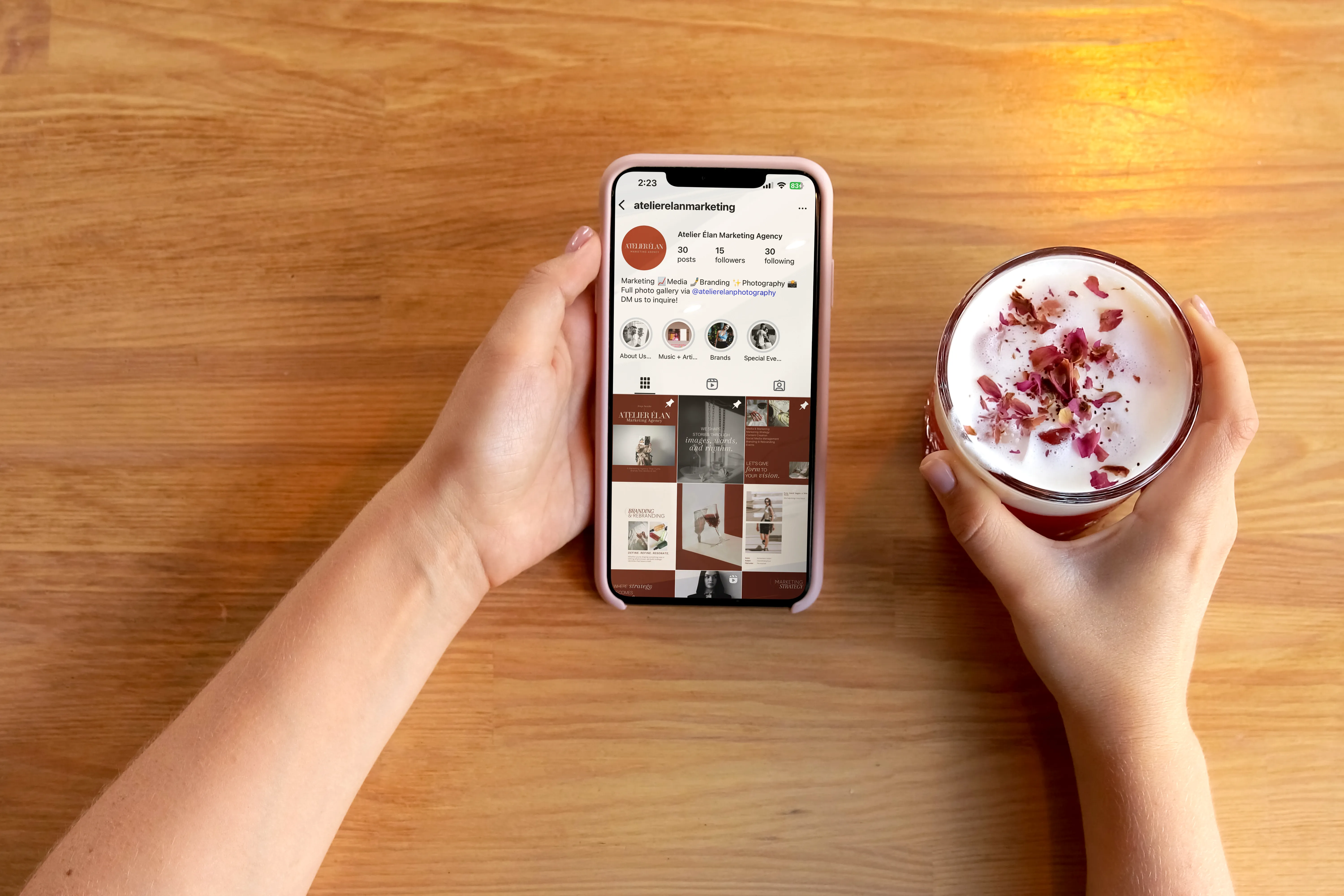 Hands holding a phone displaying Atelier Élan Marketing Agency’s Instagram feed, featuring a cohesive grid of warm rust and cream-toned posts. A rose latte sits on the wooden table beside it, creating a calm, lifestyle-inspired scene.