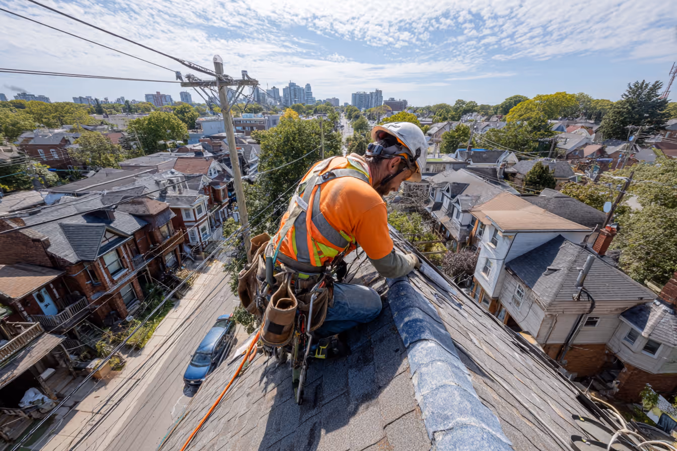 Roofer in safety harness kneeling on a sloped residential roof, preparing shingles against a backdrop of houses and tree-lined streets.