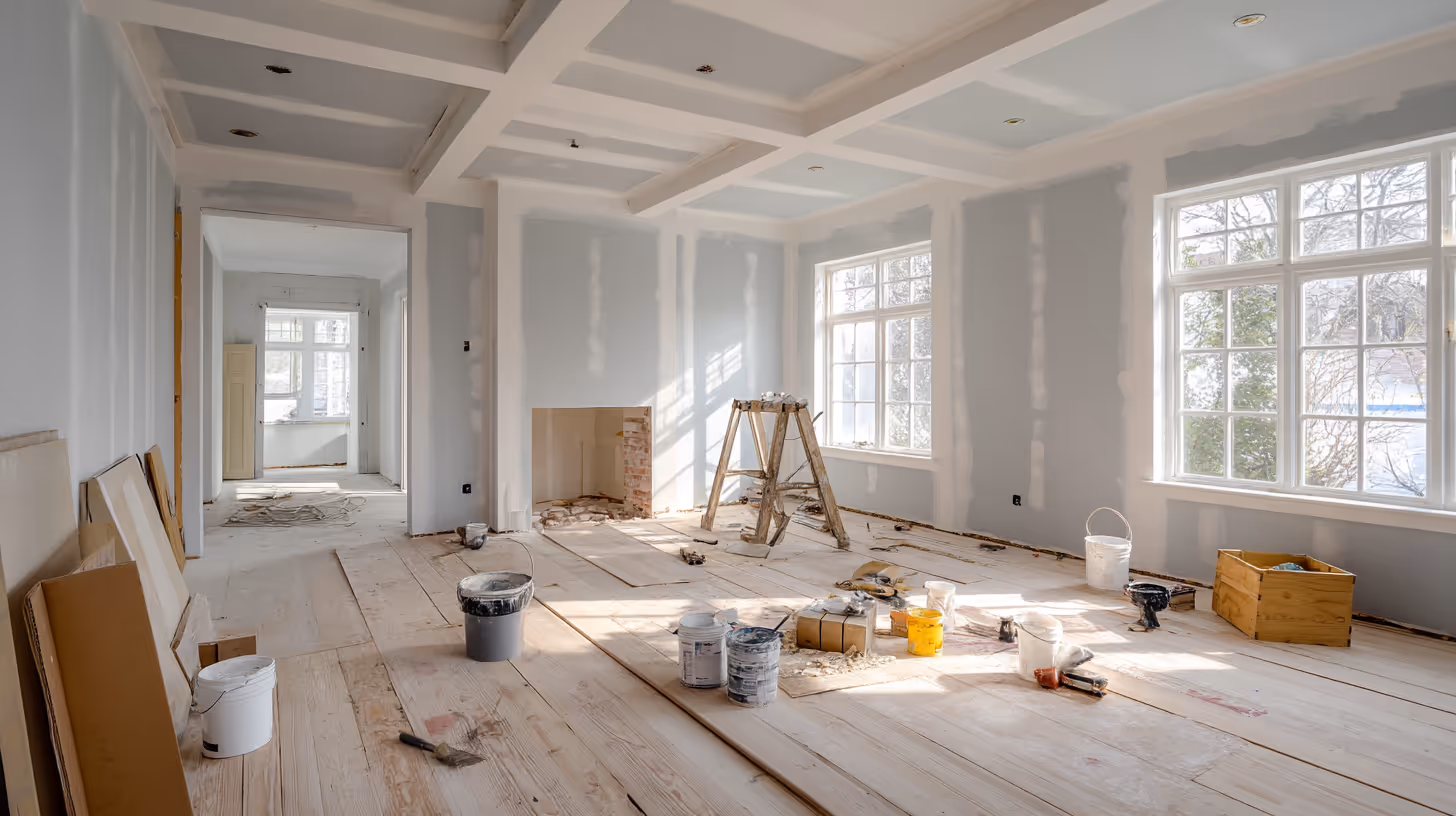 Spacious living room under renovation, with freshly installed drywall, painting supplies, and exposed wood floors awaiting finishing.