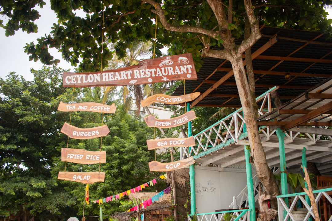 Wooden sign of Ceylon Heart Restaurant hanging under palm trees near Dewata Beach, Galle – partner restaurant of Beach Love Surf School.
