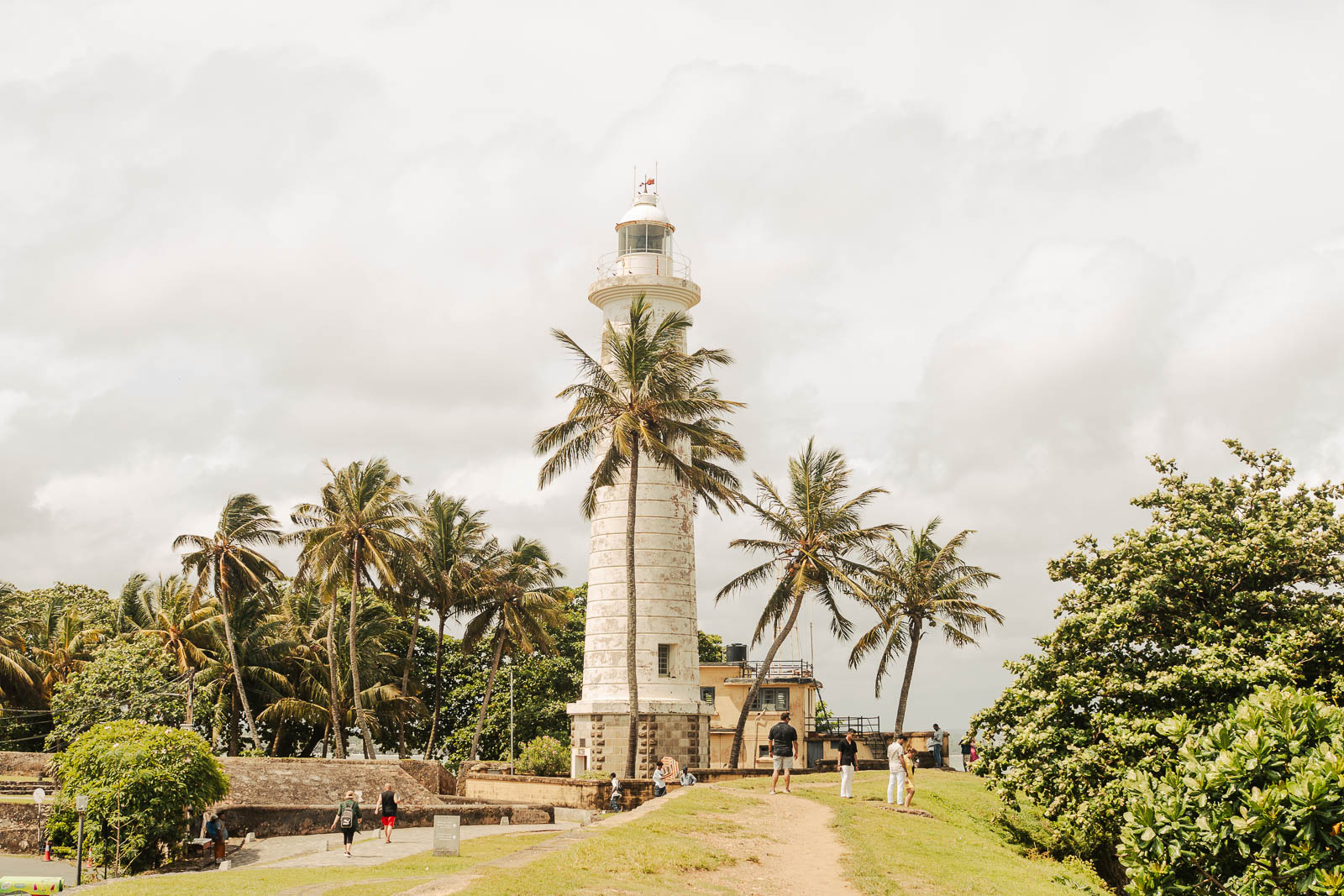 Galle Fort Lighthouse surrounded by palm trees and visitors on a sunny day in South Sri Lanka.