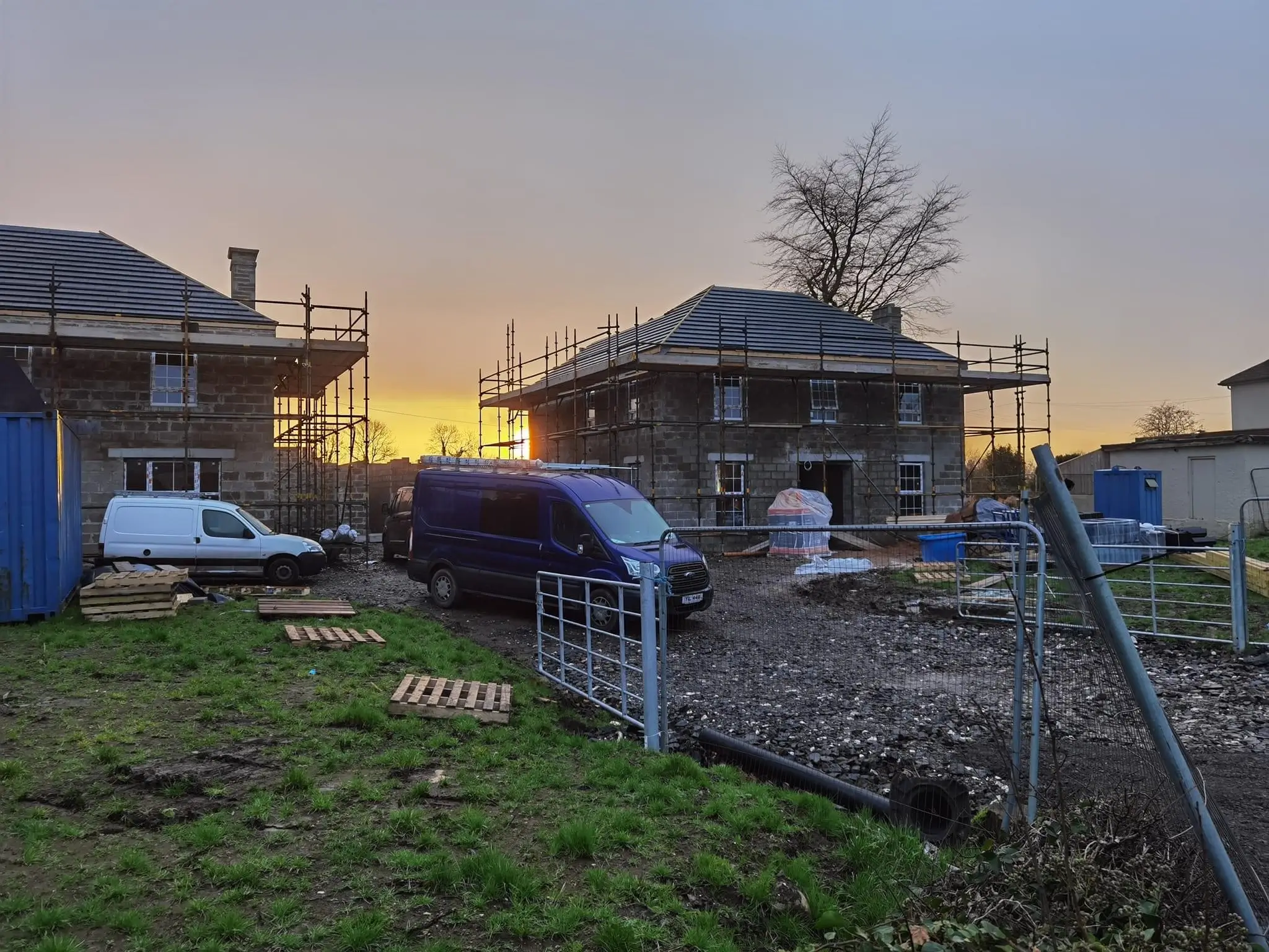 An image of a new home under construction with electrical contractors on site. The sun rises or sets gently in the background, behind the house.