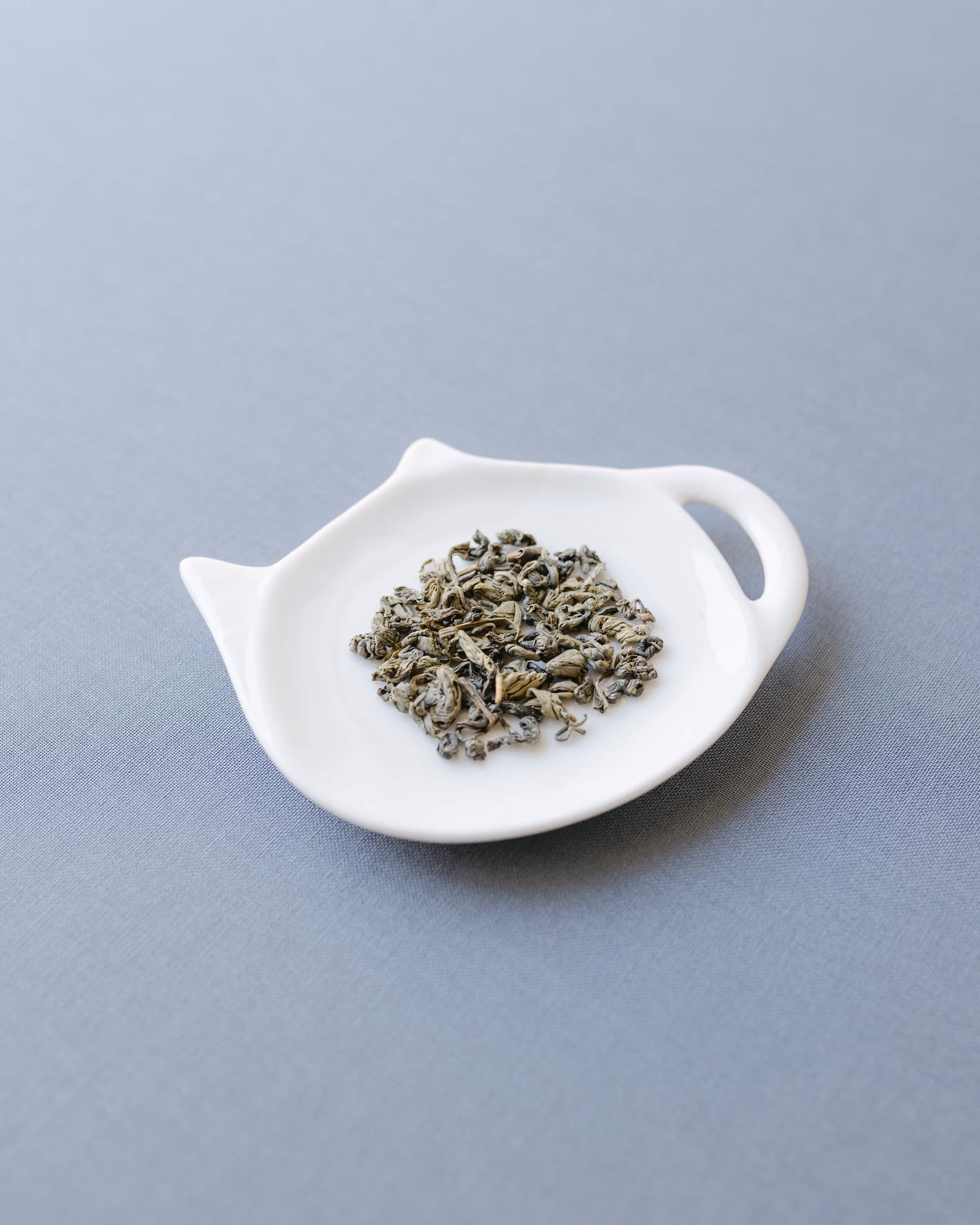 Foglie di tè sfuse su un piattino bianco a forma di teiera, dal design morbido e arrotondato.