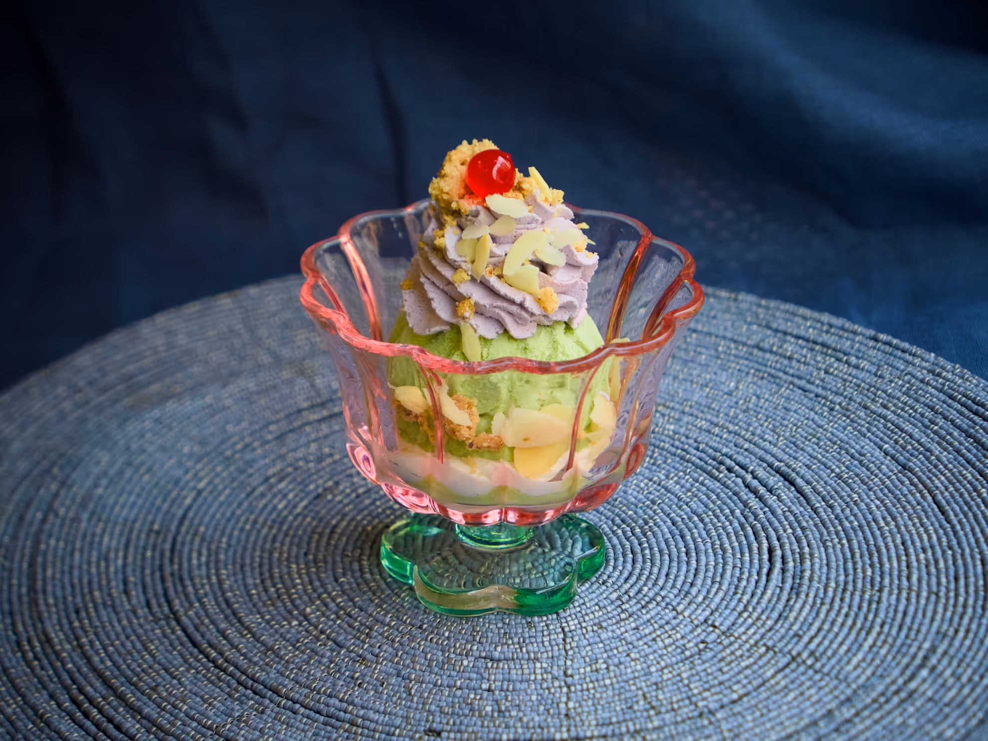 Coppa in vetro con gelato verde, panna lilla, scaglie di mandorla e una ciliegia rossa in cima.