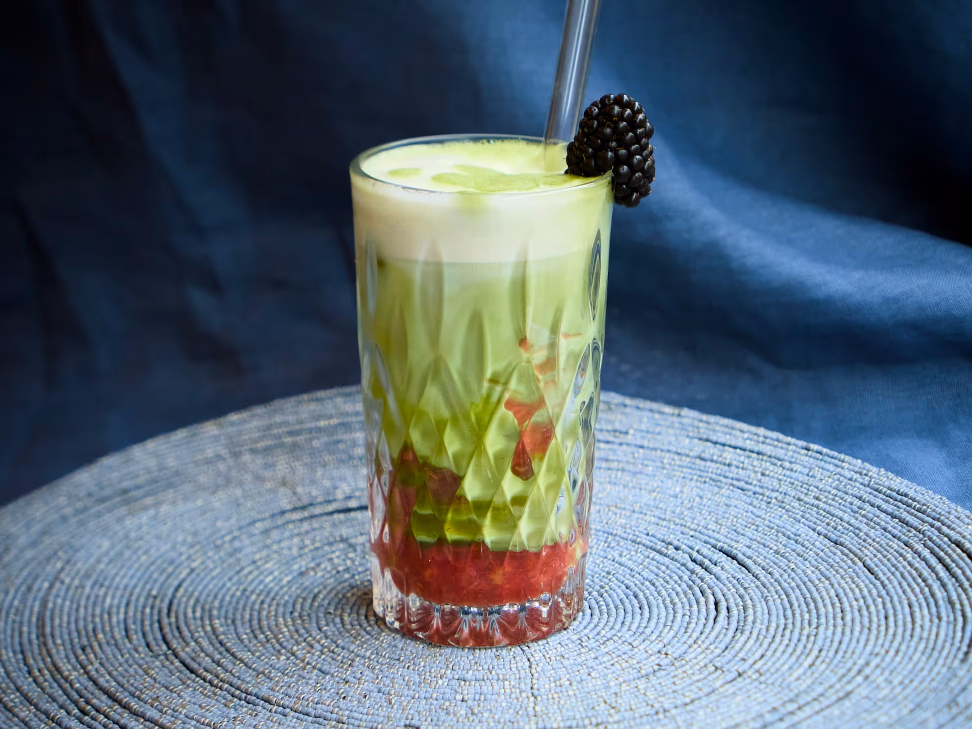 Bevanda a strati con base rossa, liquido verde e schiuma bianca, guarnita con una mora e cannuccia in vetro.