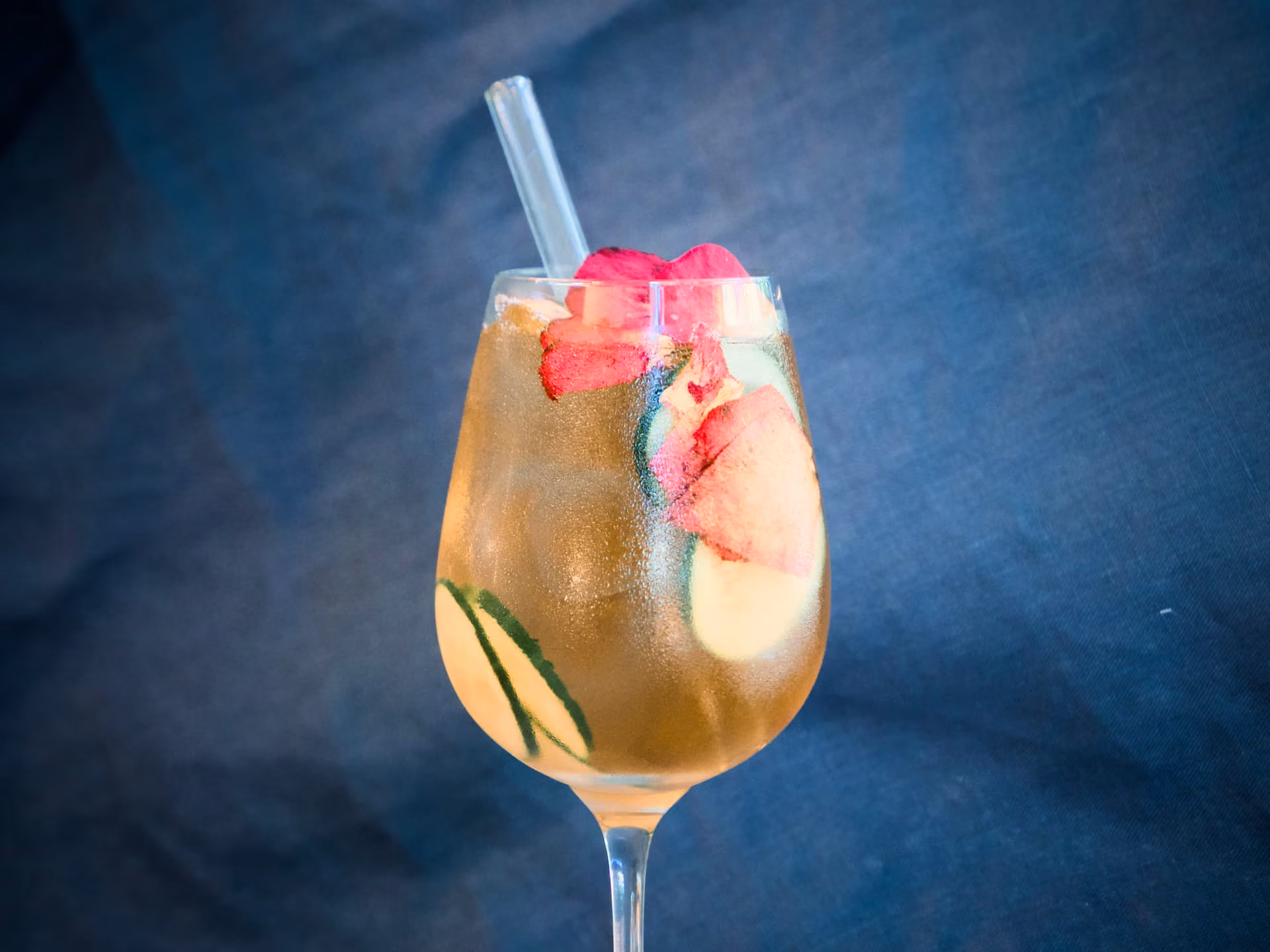 Tè bianco freddo in un calice con petali di rosa, fettine di cetriolo, ghiaccio e cannuccia di vetro.