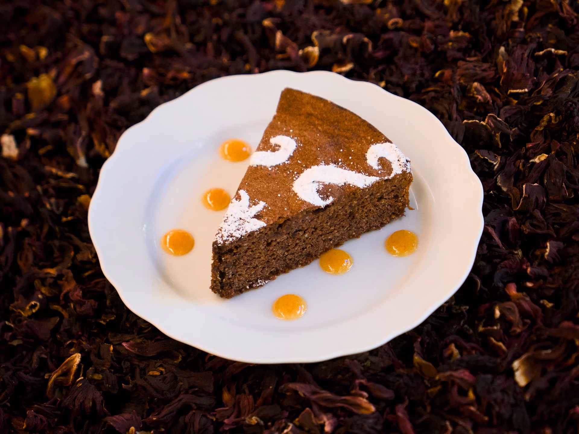 Fetta di torta scura decorata con zucchero a velo e gocce di salsa al mandarino, su piattino bianco.