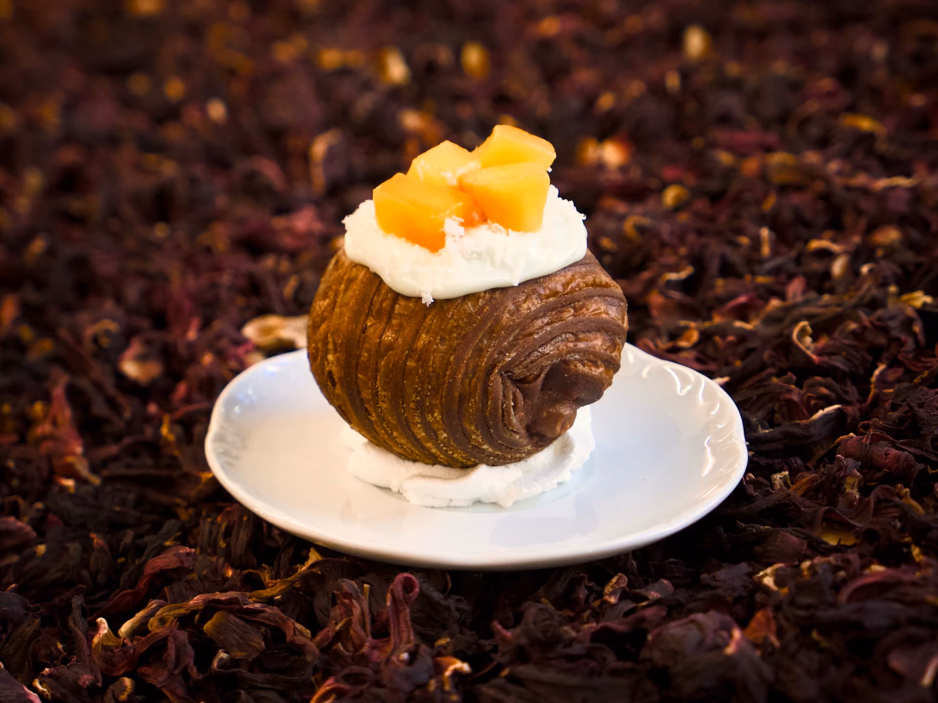 Croissant sfogliato con crema bianca, cocco rapé e cubetti di frutta arancione, su piattino bianco.