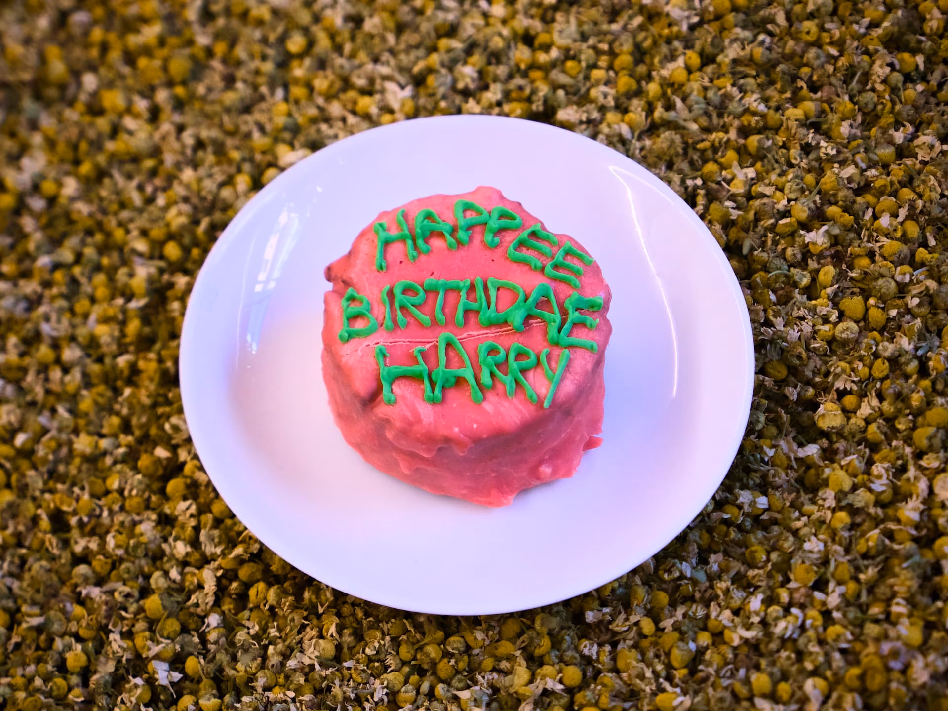 Tortina rosa con la scritta verde «HAPPEE BIRTHDAE HARRY» su un piatto bianco, sopra fiori di camomilla essiccati.
