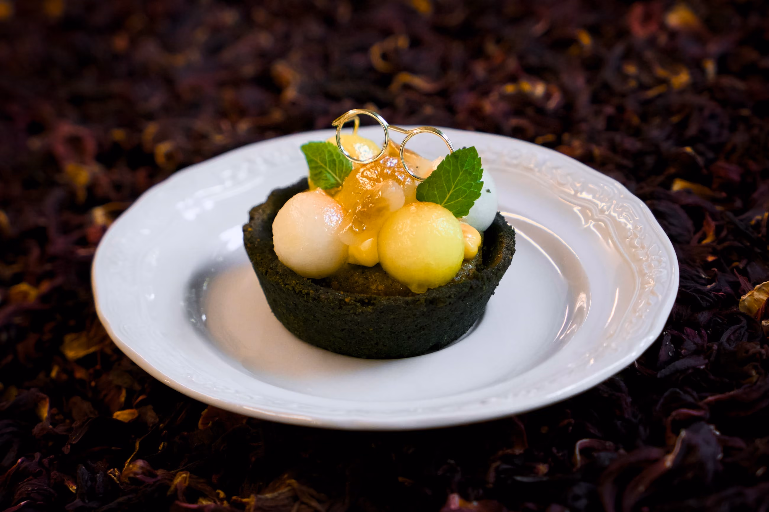 Crostatina scura con palline di frutta fresca, frangipane al tè matcha e foglie di menta, decorata con un paio di occhiali tondi dorati.