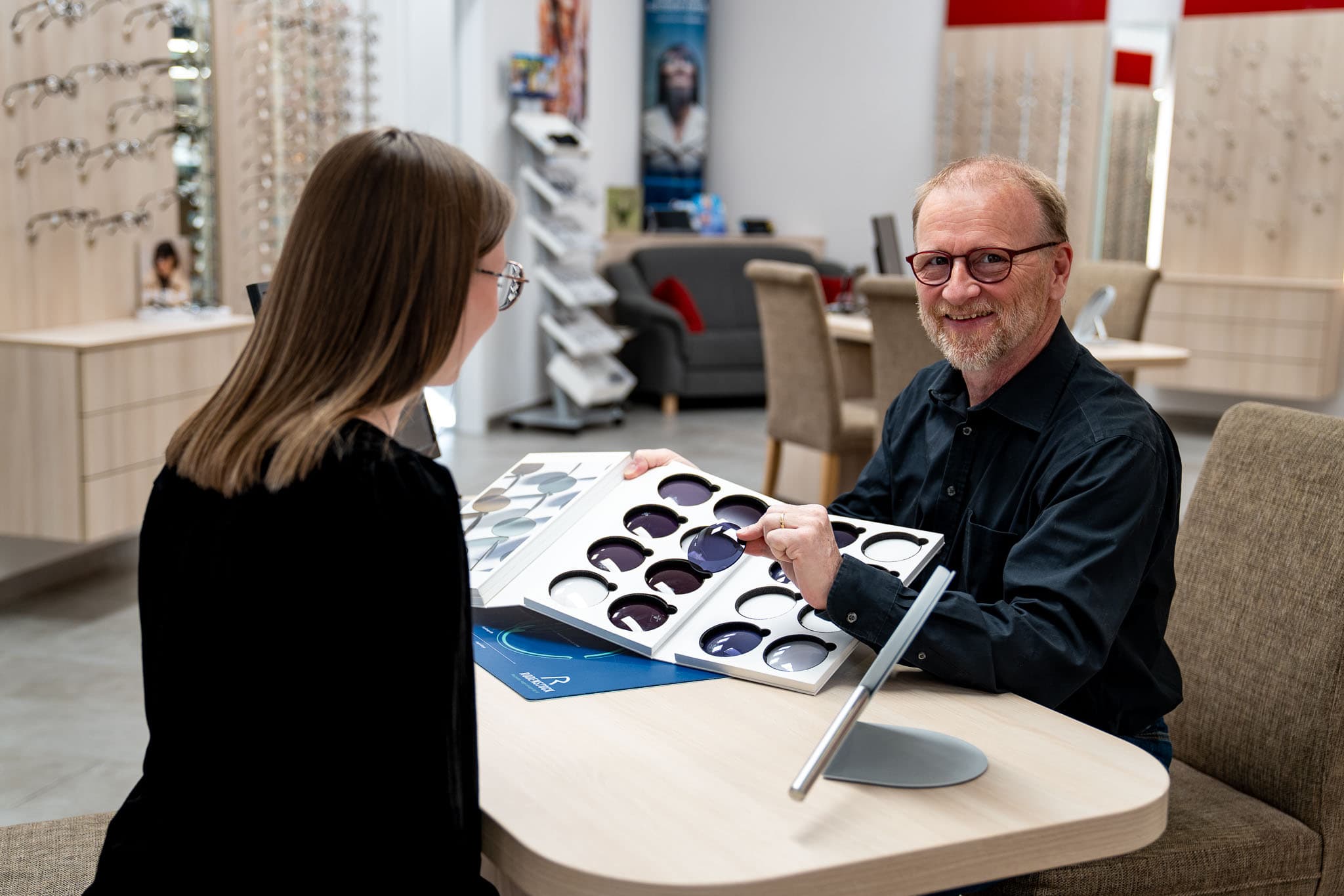 Zwei Personen, eine männliche und eine weibliche Person. Die männliche Person zeigt der weiblichen Person verschiedenen Brillengläser