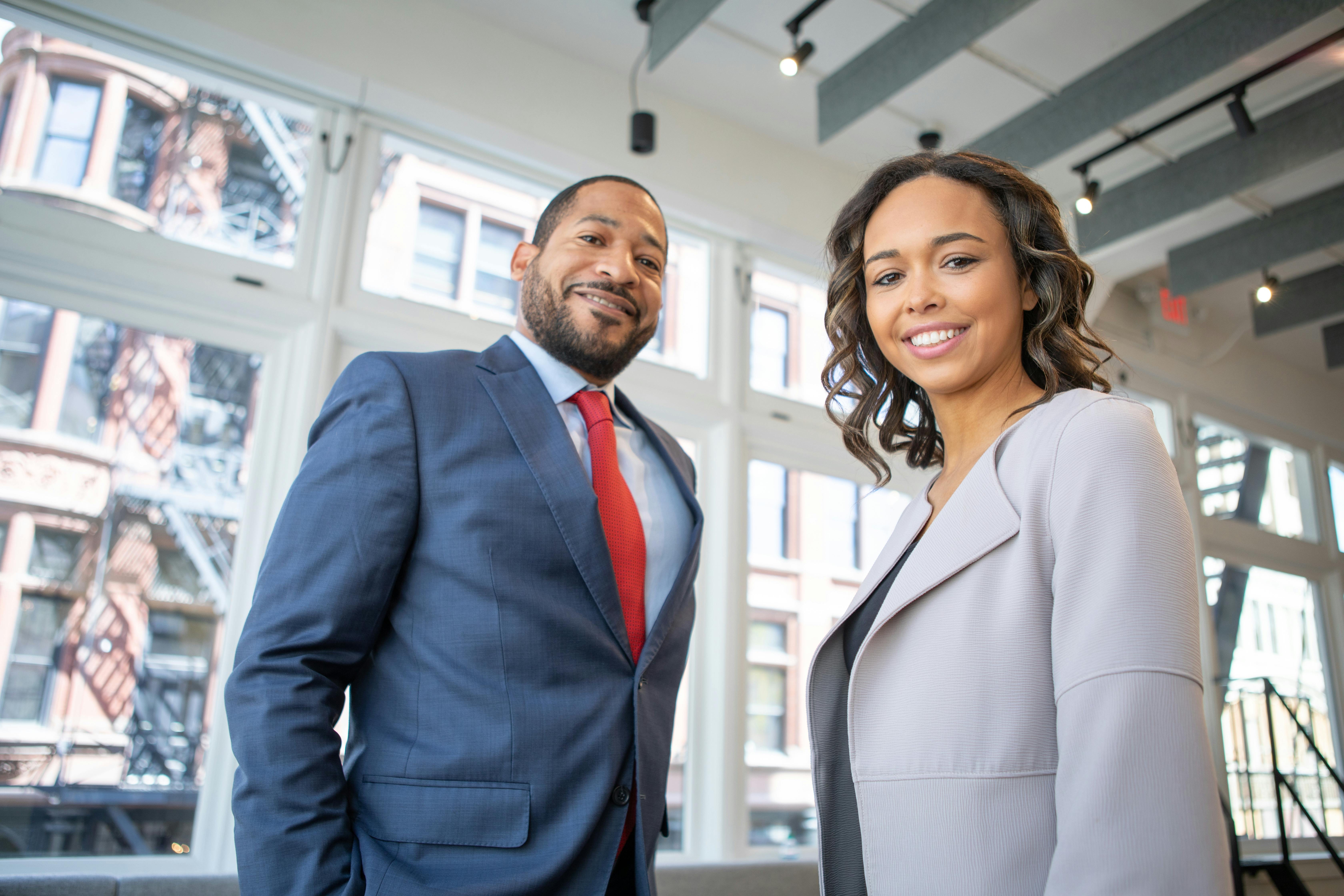 Three legal professionals sit at a wooden conference table with law bookshelves in the background. The central figure, a smiling Black man in a dark suit and red tie, is flanked by two women in professional attire, creating an inviting and collaborative law firm atmosphere.
