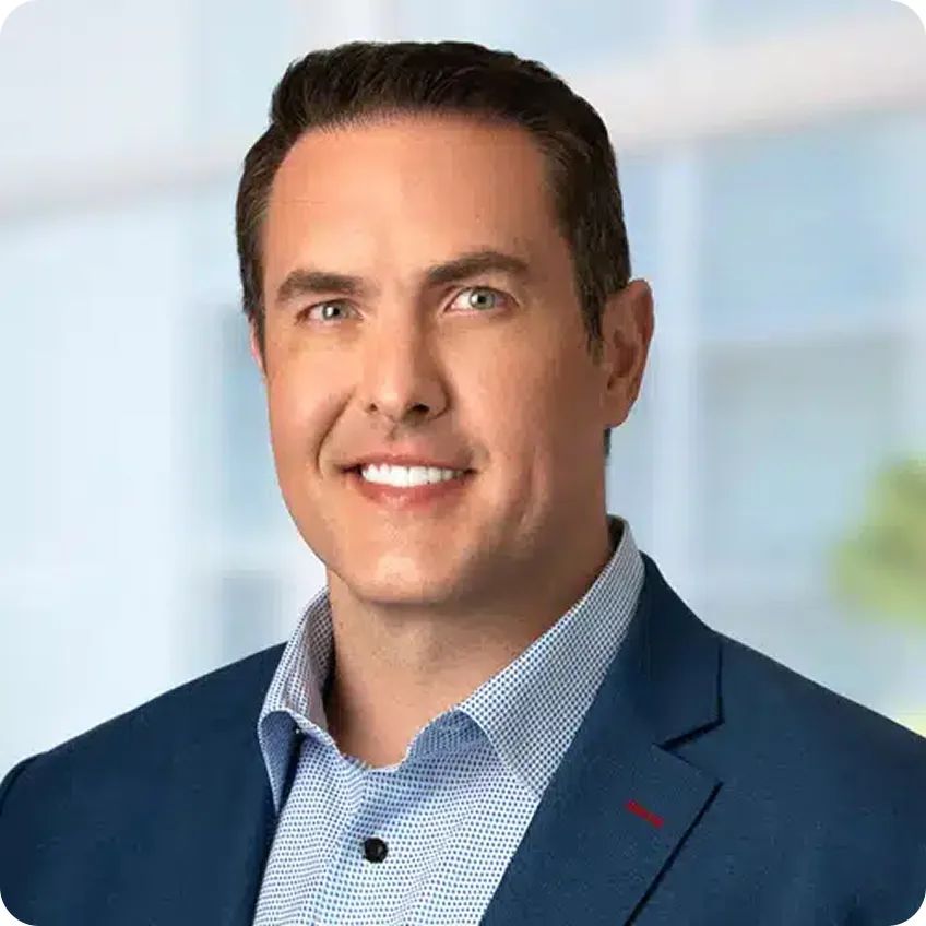Joe Welu in a blue suit jacket and patterned shirt, smiling in a corporate office setting