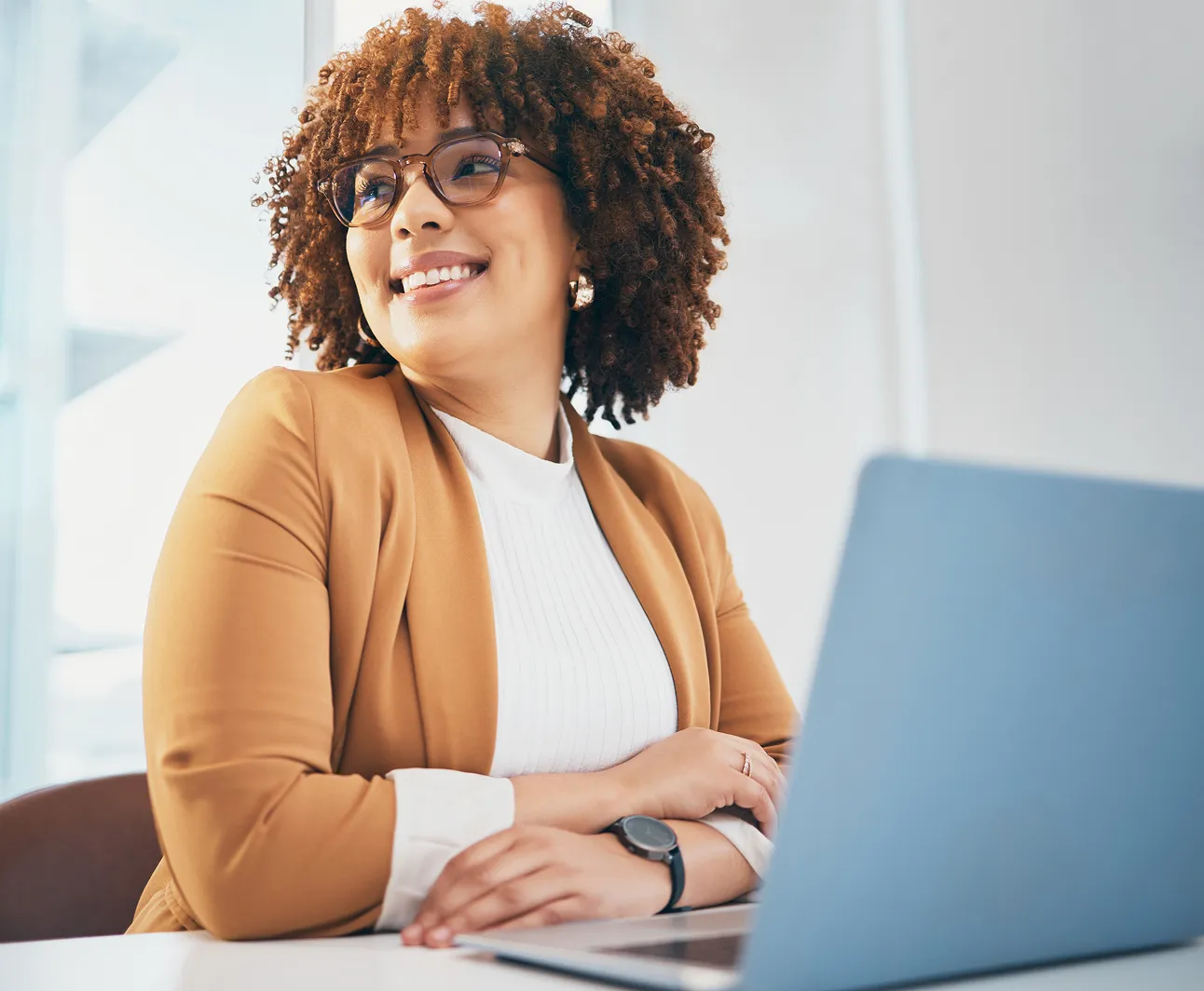 Smiling professional woman with curly hair wearing glasses and a tan blazer, sitting at a desk with a laptop