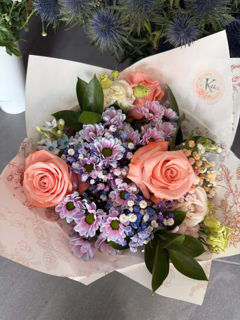 Fresh flower arrangement featuring pastel roses and lavender chrysanthemums, wrapped in decorative paper with Kikkis logo.