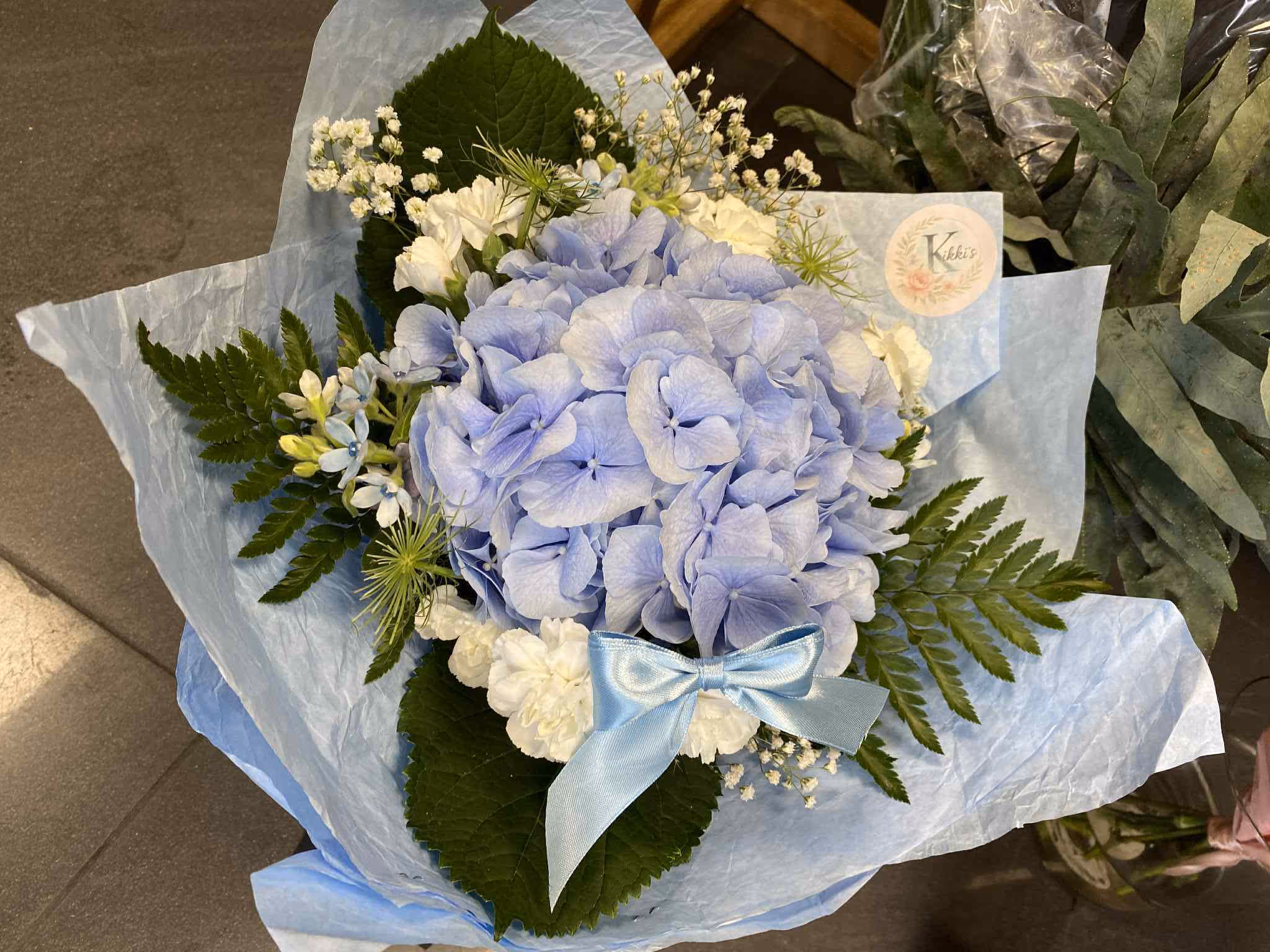 Uroczy bukiet z jasnoniebieską hortensją, białymi dodatkami, paprocią i ozdobną wstążką – zapakowany z etykietą Kikki’s.