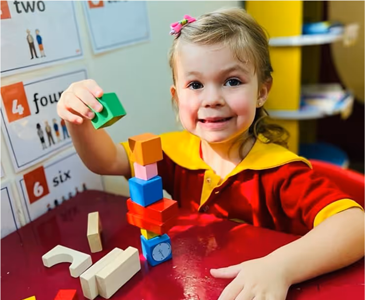 Smiling young girl stacking colorful wooden blocks at a red table in a classroom with number posters on the wall.