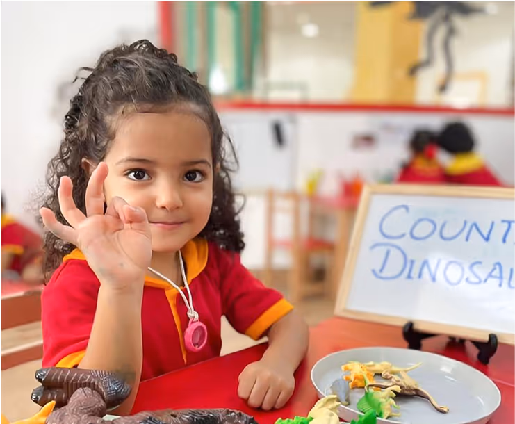 Young girl in red outfit sitting at a table with toy dinosaurs, holding up an OK hand gesture.
