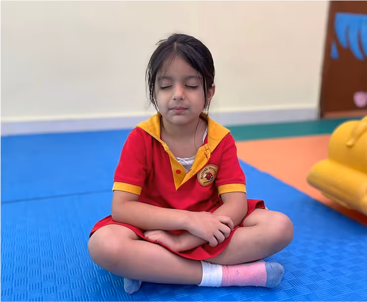 Young girl in a red and yellow school uniform sitting cross-legged on a blue mat with eyes closed and hands resting on her lap.