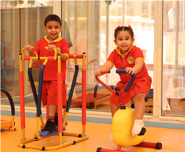 Two young children in red school uniforms playing on indoor exercise equipment in a brightly lit room.