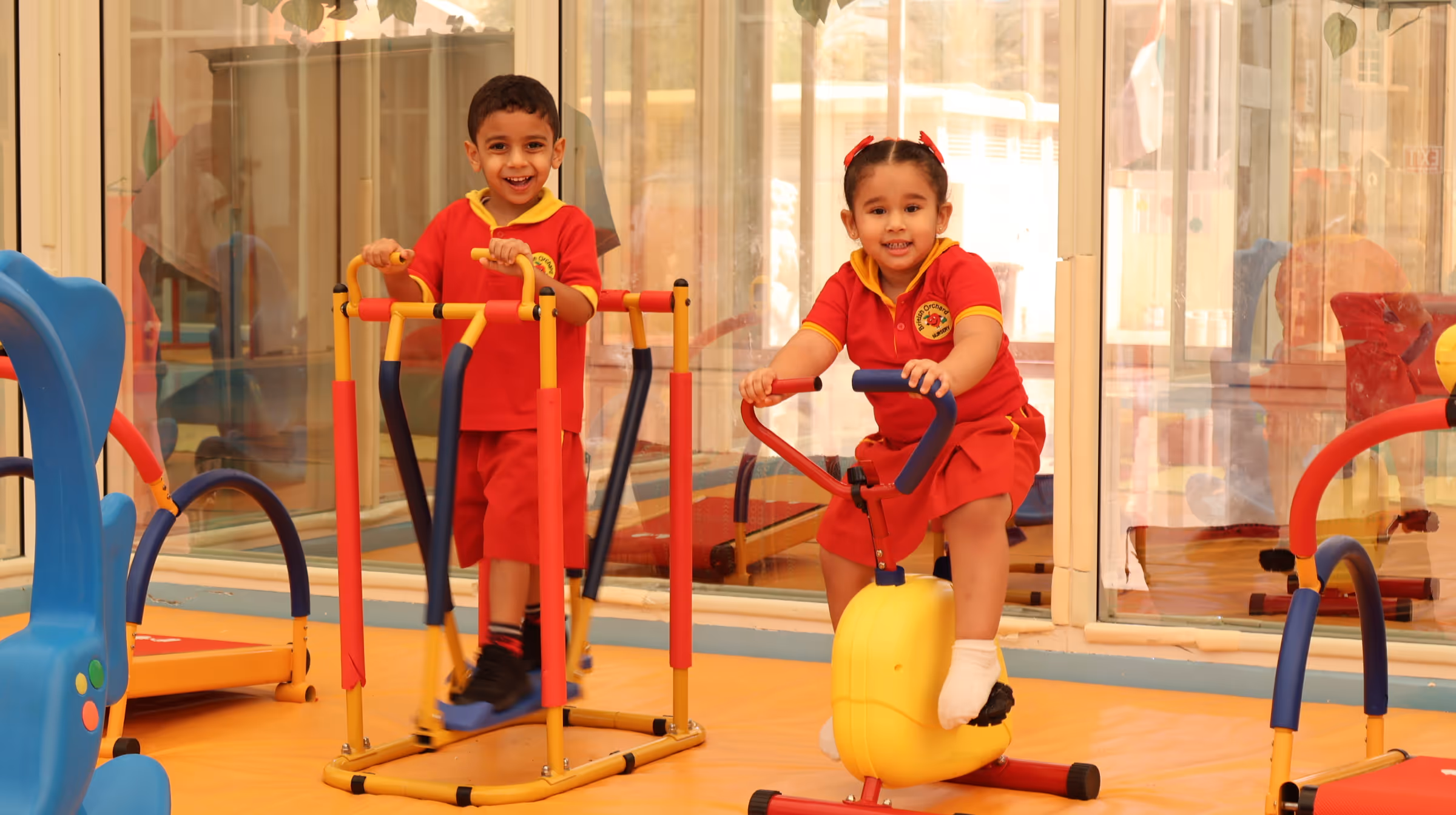 Two young children in red and yellow uniforms playing with indoor exercise equipment in a bright room.