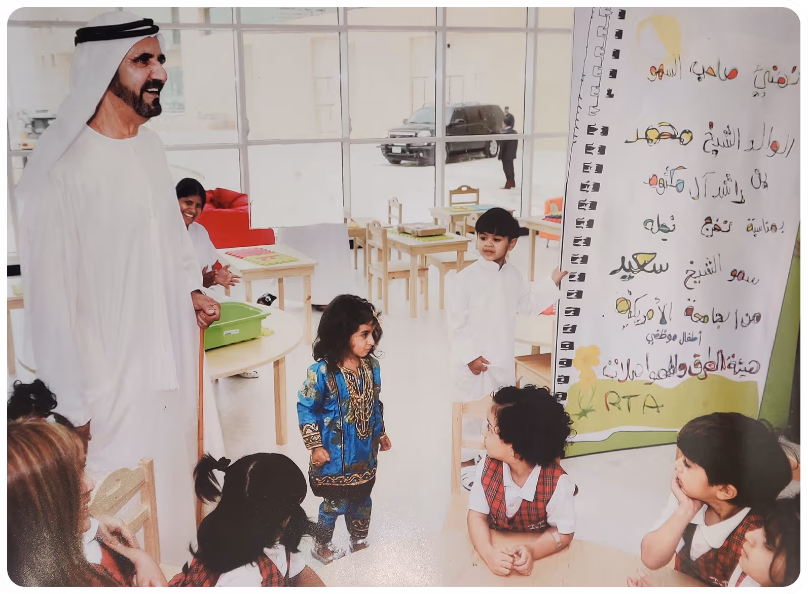 Children in a classroom with a man in traditional Emirati clothing holding a cane, standing beside a large board with Arabic writing.