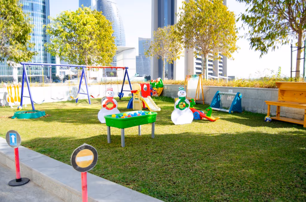 Outdoor children's playground with swings, slides, toy snowmen, a ball pit, and a small soccer goal on green grass under sunlight.