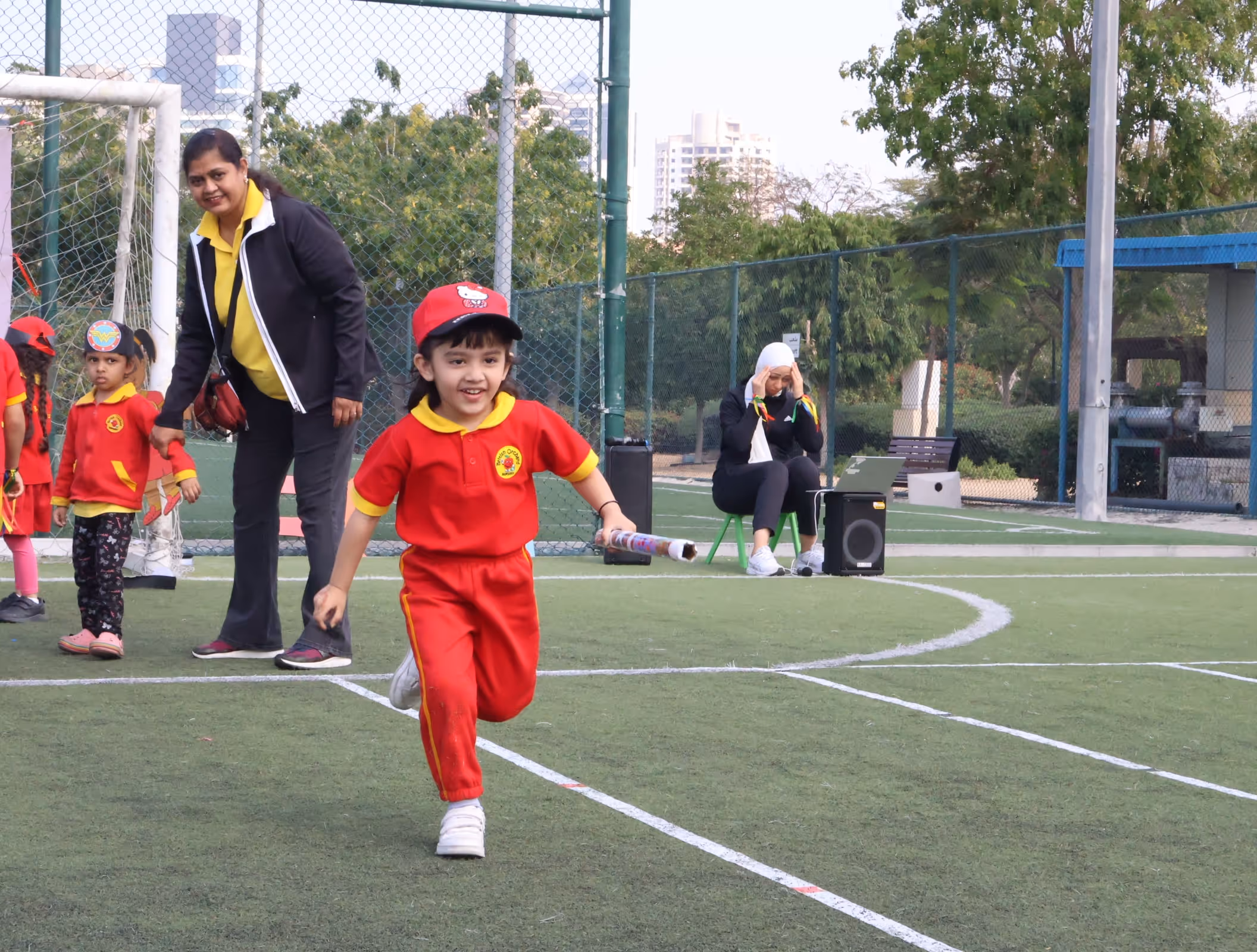 Young child in red sportswear running on a marked artificial turf field holding a rolled paper, with adults and other children in the background.