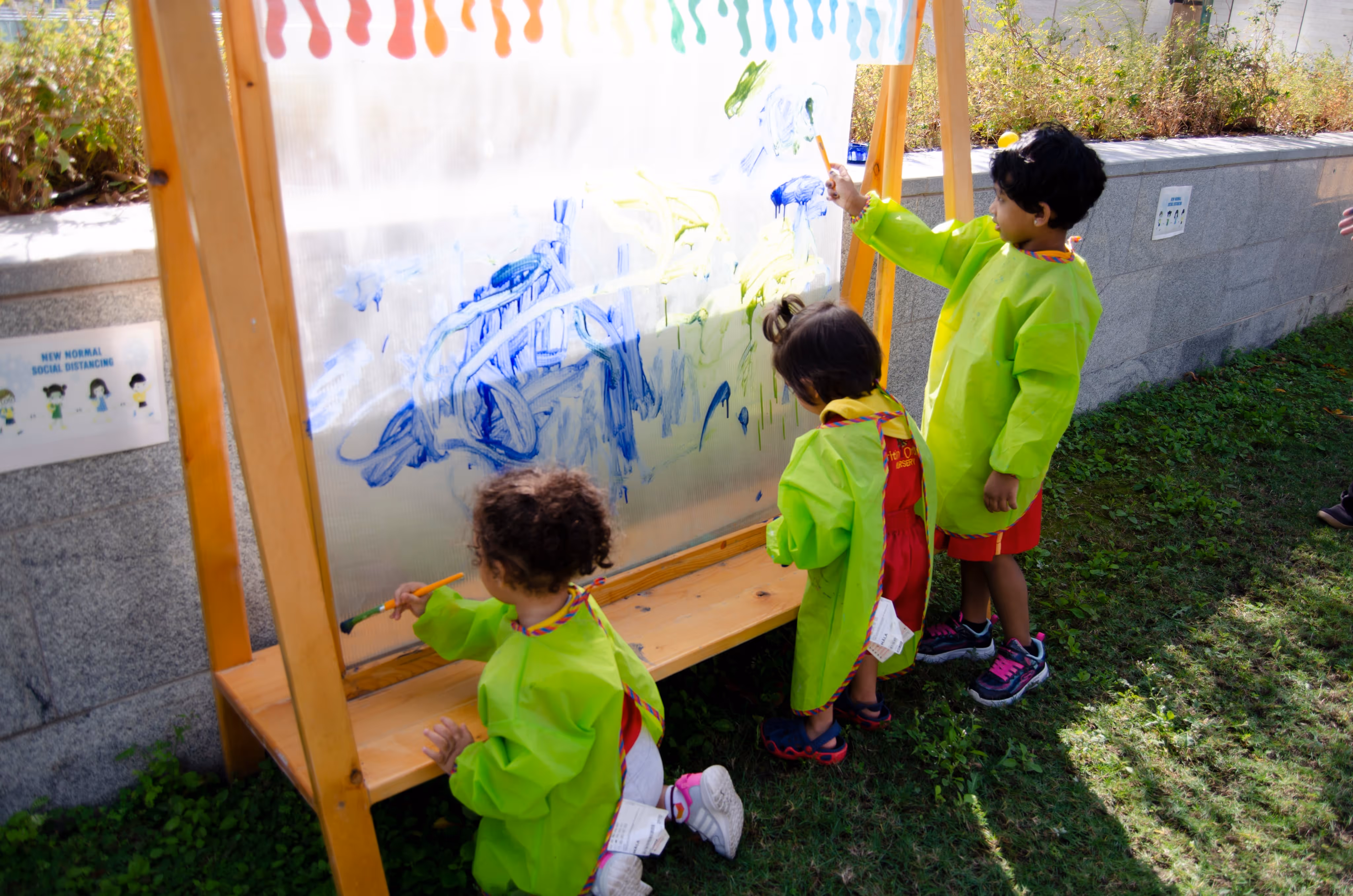 Three children in green smocks painting on a transparent easel outdoors near a stone wall.