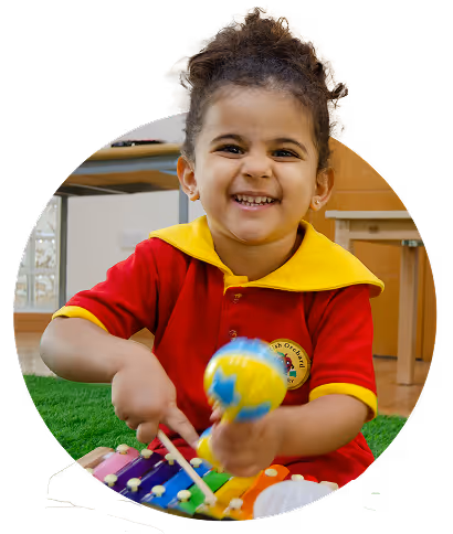 Smiling young child playing colorful xylophone and holding a globe-shaped maraca in a classroom.