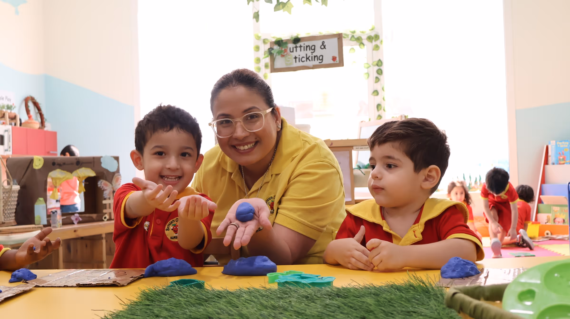 Teacher and two young children playing with blue clay on a yellow table in a colorful classroom.