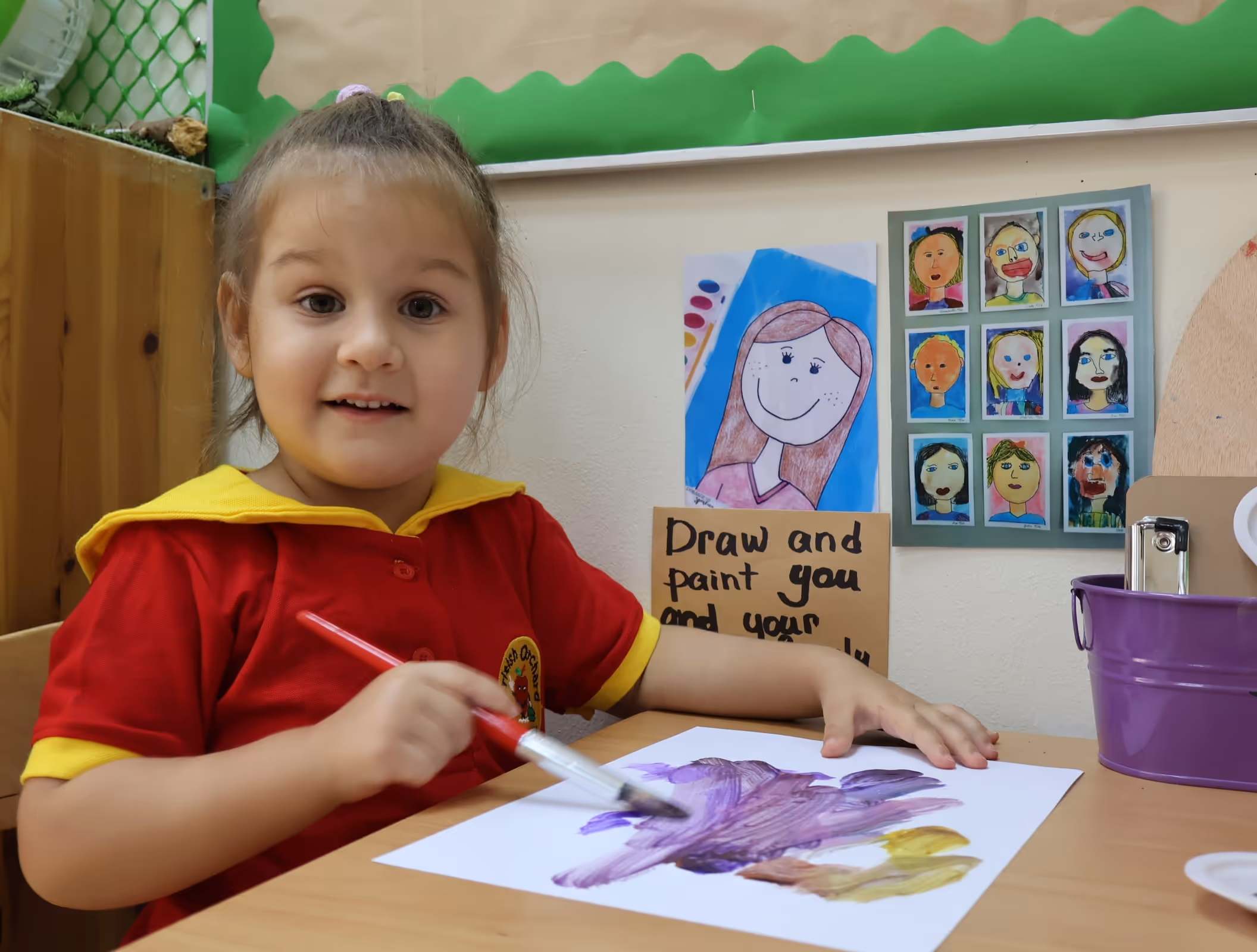 A young girl in a red and yellow shirt painting with a brush on white paper at a table in a classroom with children's artwork on the wall behind her.