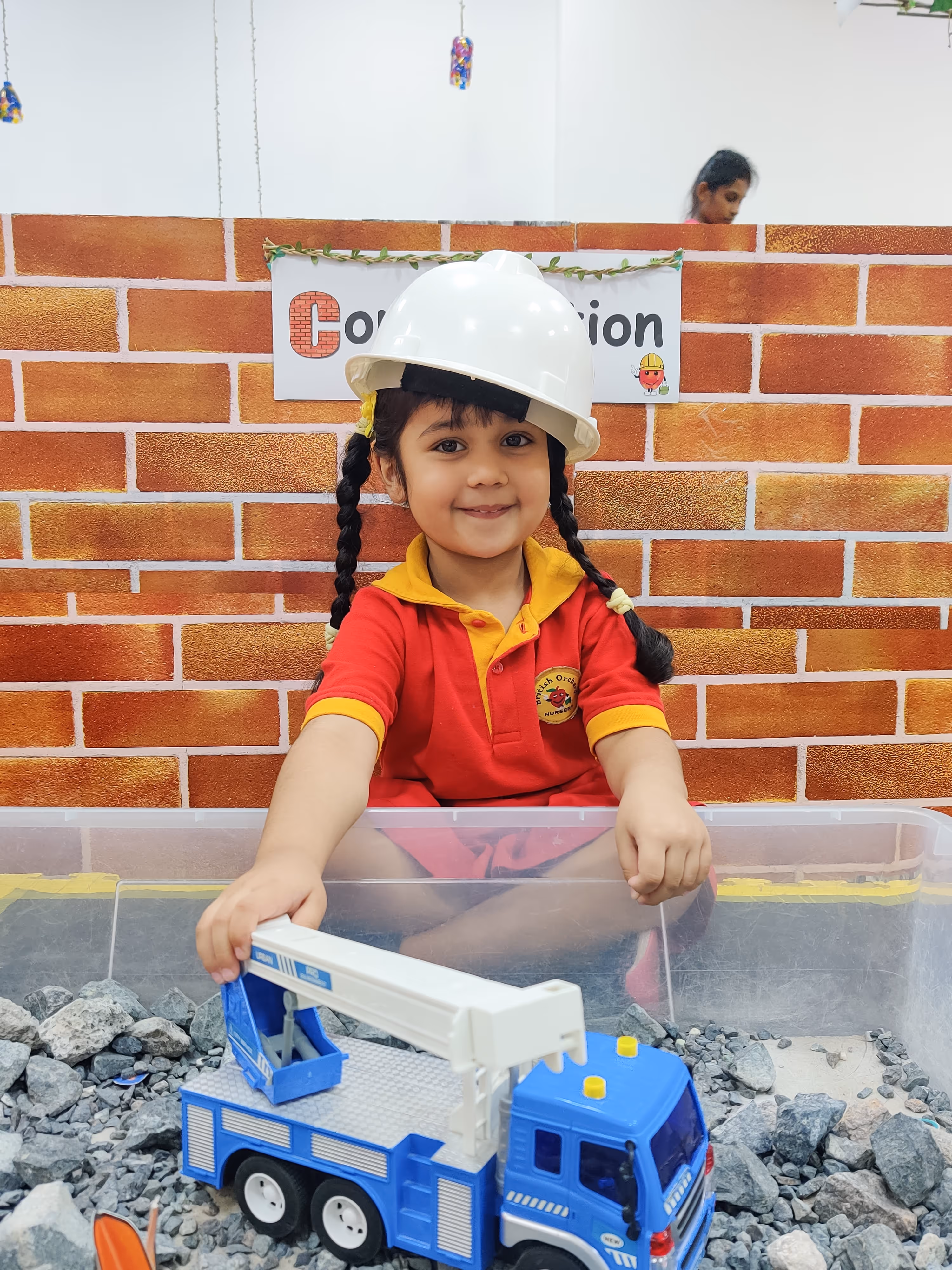 Young girl wearing a white construction helmet playing with a blue toy crane truck in a play area with rocks and a brick wall backdrop.