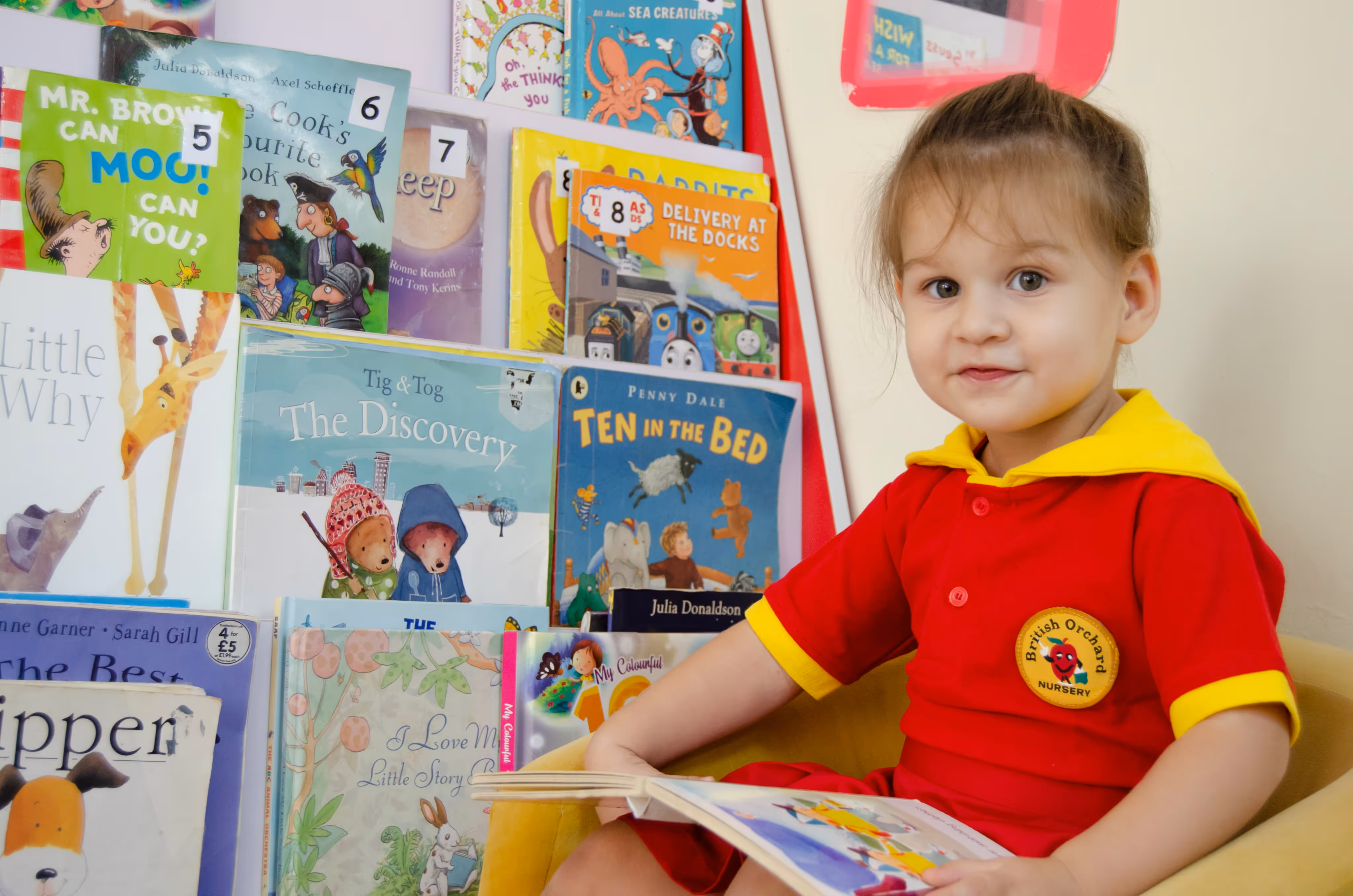 A young child in a red and yellow British Orchard Nursery uniform sitting in a yellow chair, reading a book with a colorful collection of children's books displayed behind.