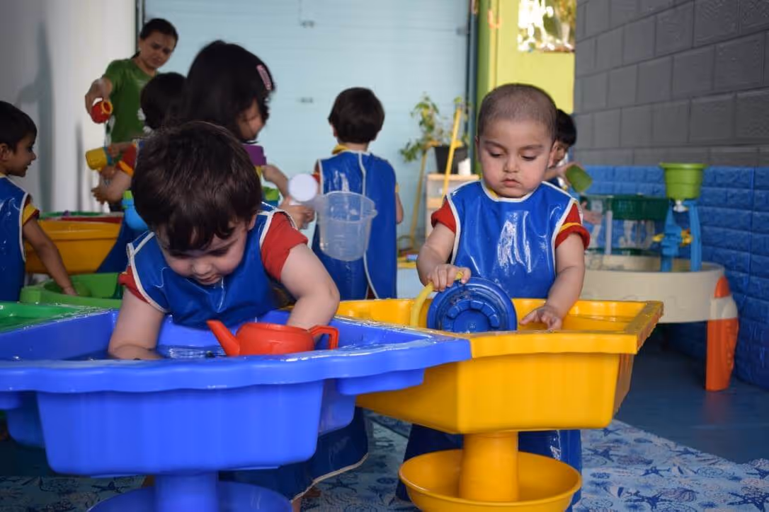 Young children playing with water tables, wearing blue aprons, engaged in sensory water play indoors.