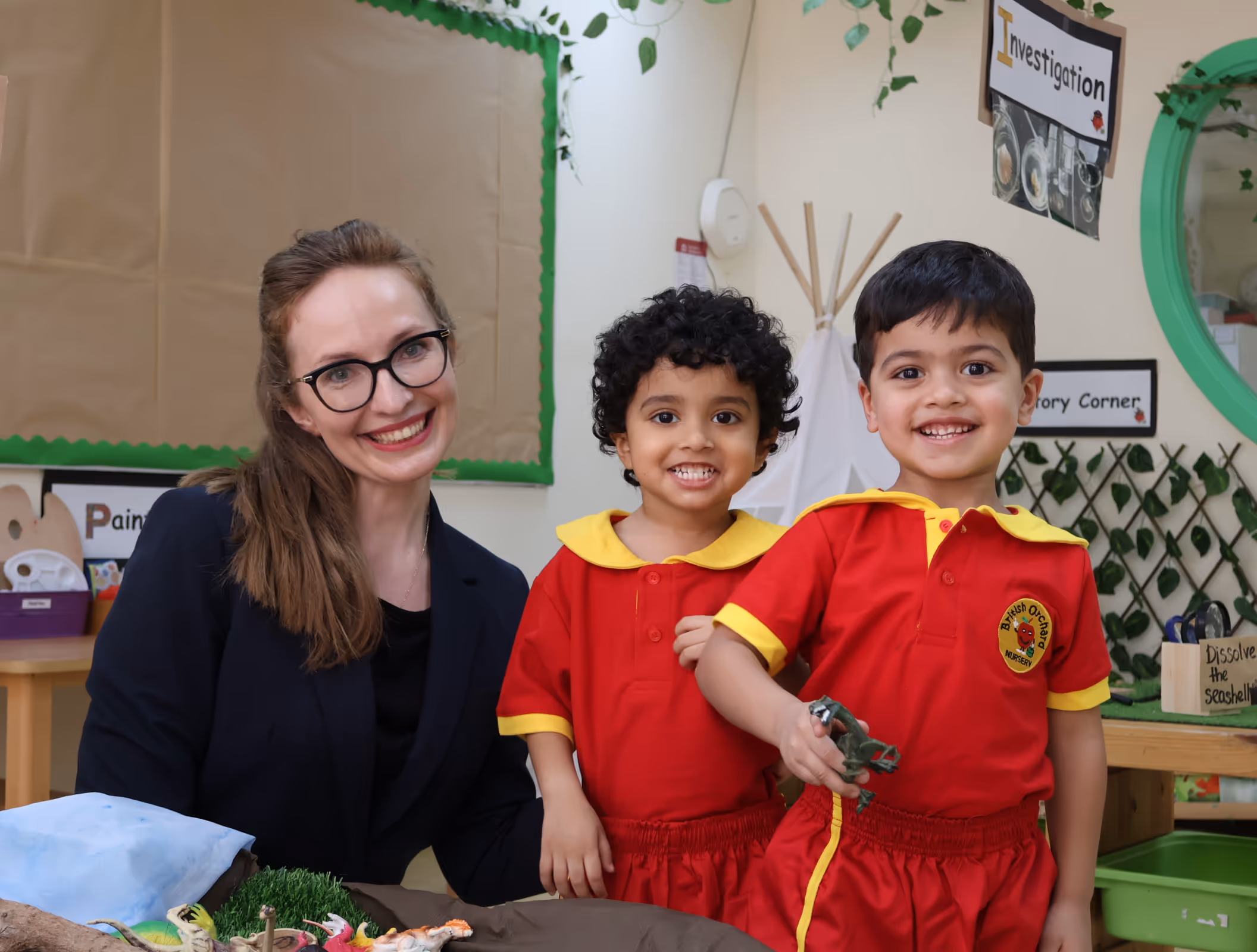 Smiling teacher with glasses poses with two young children in red and yellow nursery uniforms holding toy dinosaurs in a classroom.