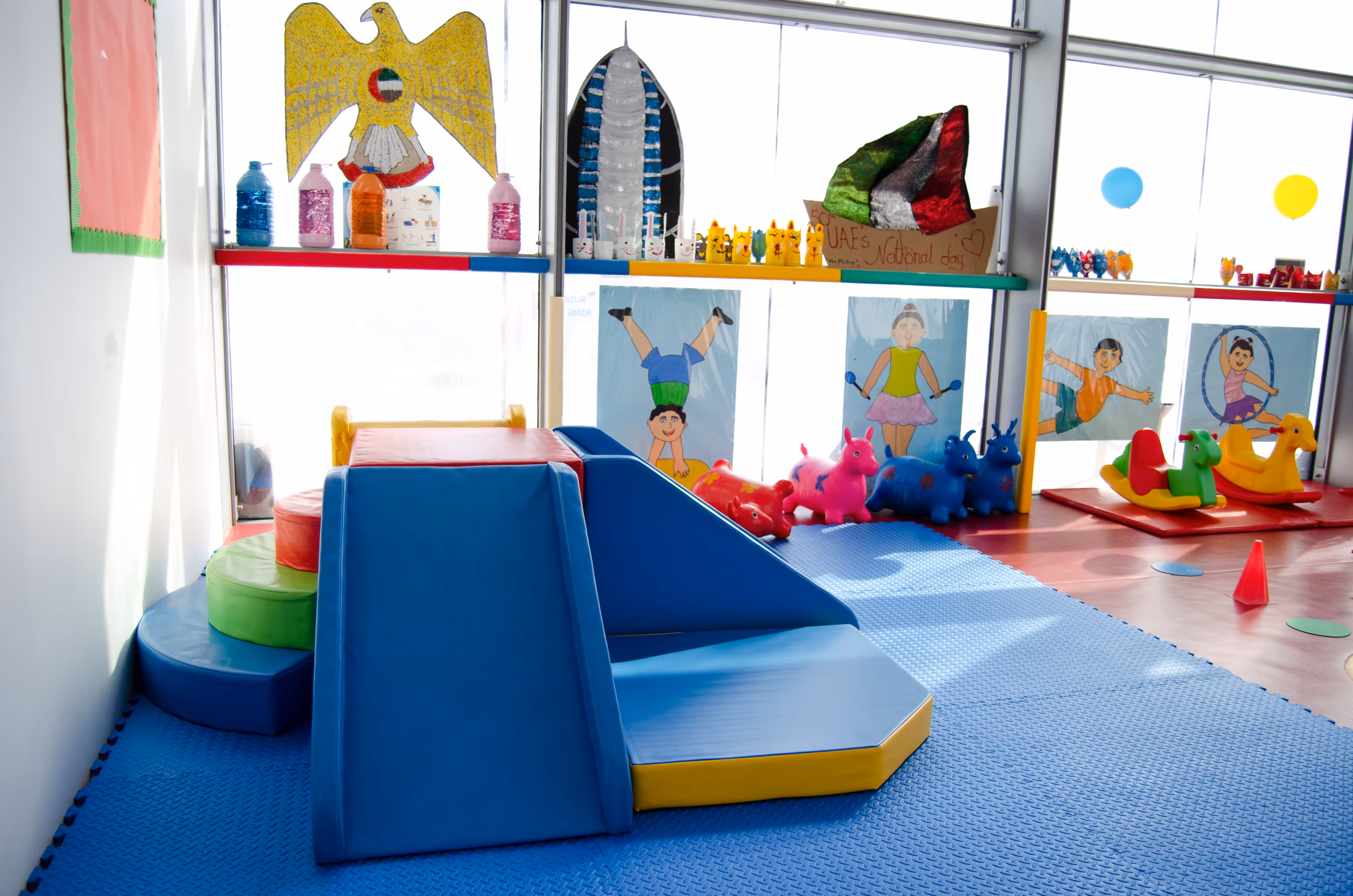 Bright indoor children's play area with soft climbing shapes, colorful ride-on toys, and wall decorations celebrating UAE National Day.
