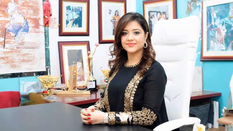 Woman in black outfit with gold embroidery sitting at a desk in an office with framed pictures and trophies behind her.