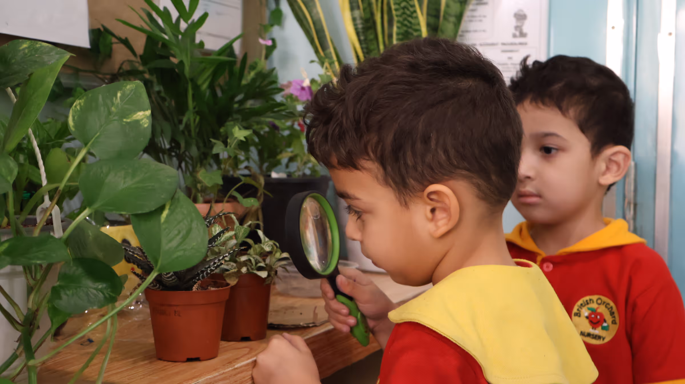 Two young boys in red and yellow uniforms examining potted plants on a table, one using a green magnifying glass.