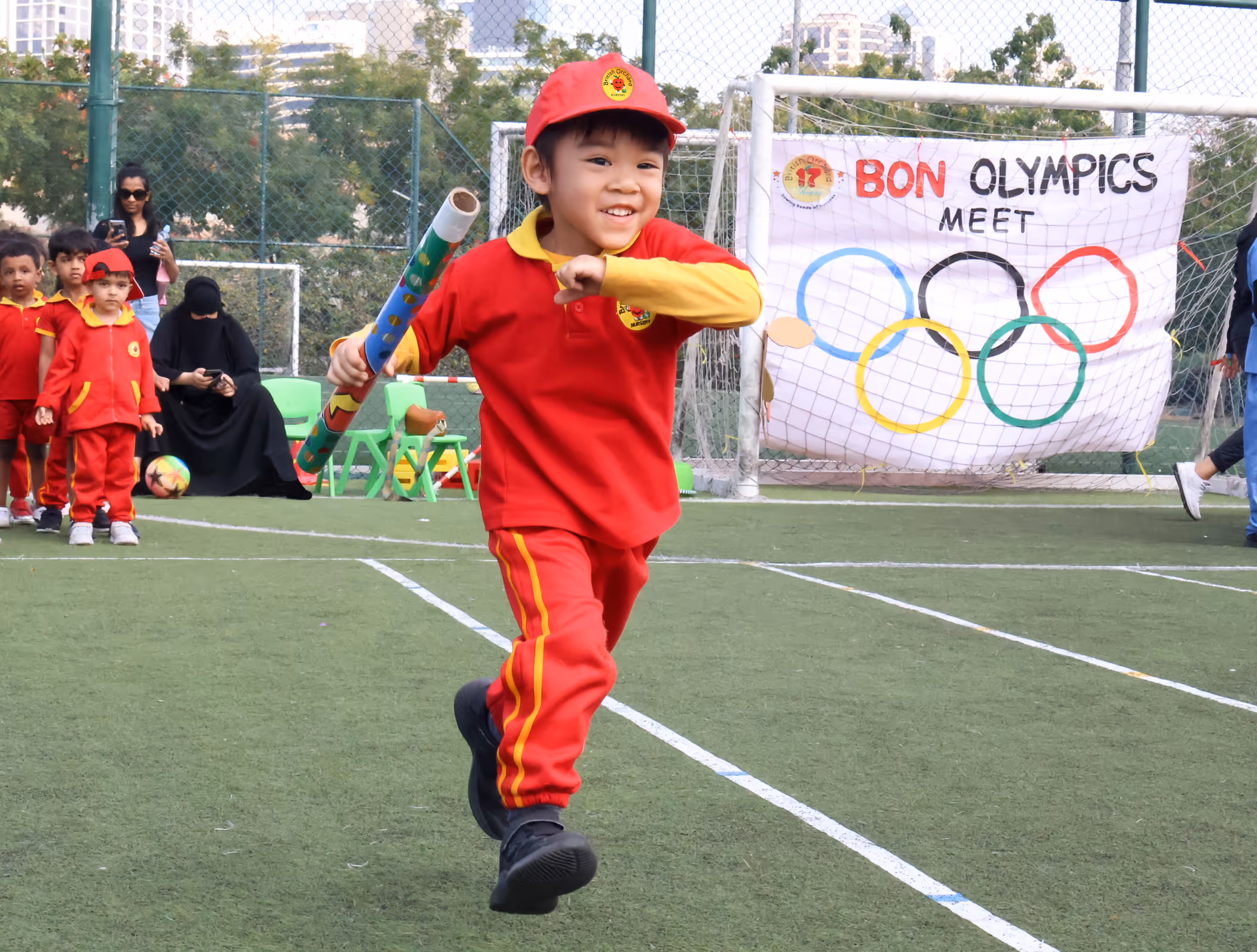 Young child in red sports attire running on a field holding a decorated baton during a children's Olympics event.