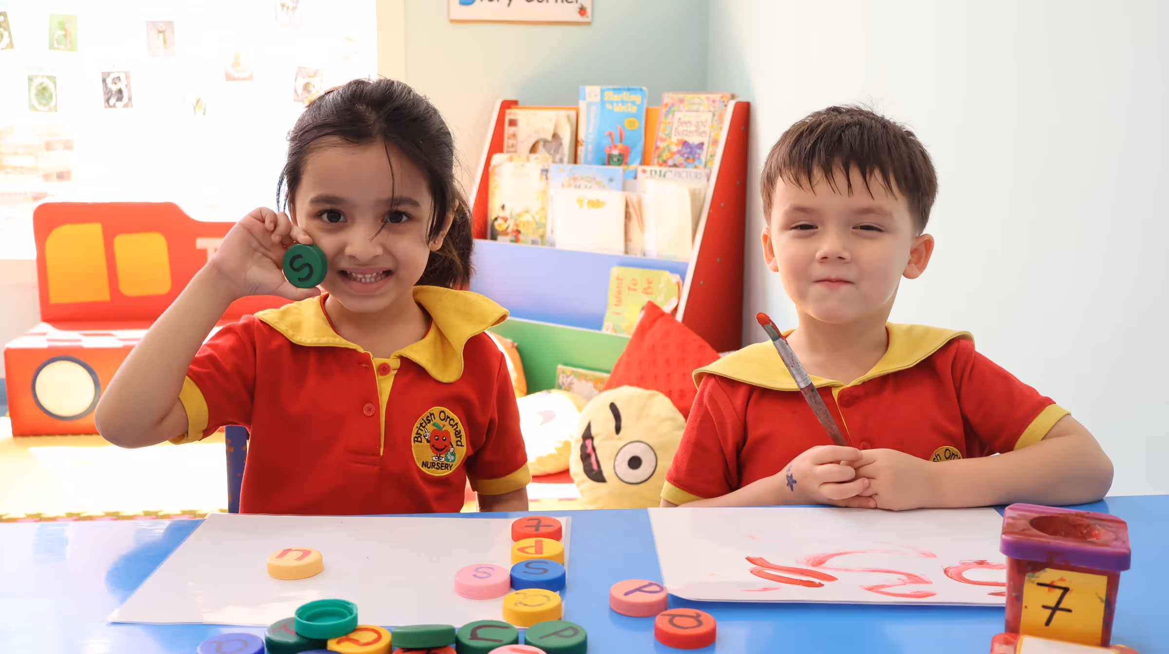 Two children in red and yellow uniforms sitting at a table with colorful alphabet and number discs, one girl smiling and holding a green disc with the letter S, and a boy holding a paintbrush with red paint on paper.